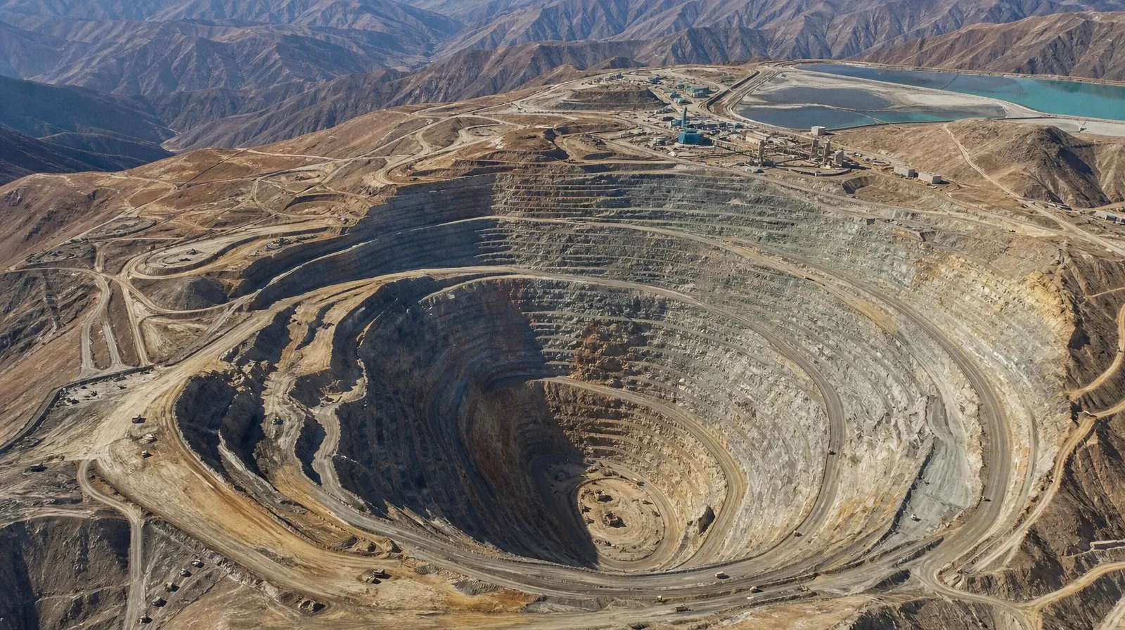 Vista aérea de un tajo abierto de gran escala labrado en bancos concéntricos sobre una cordillera árida, con la planta de procesos y la poza de relaves en el borde superior